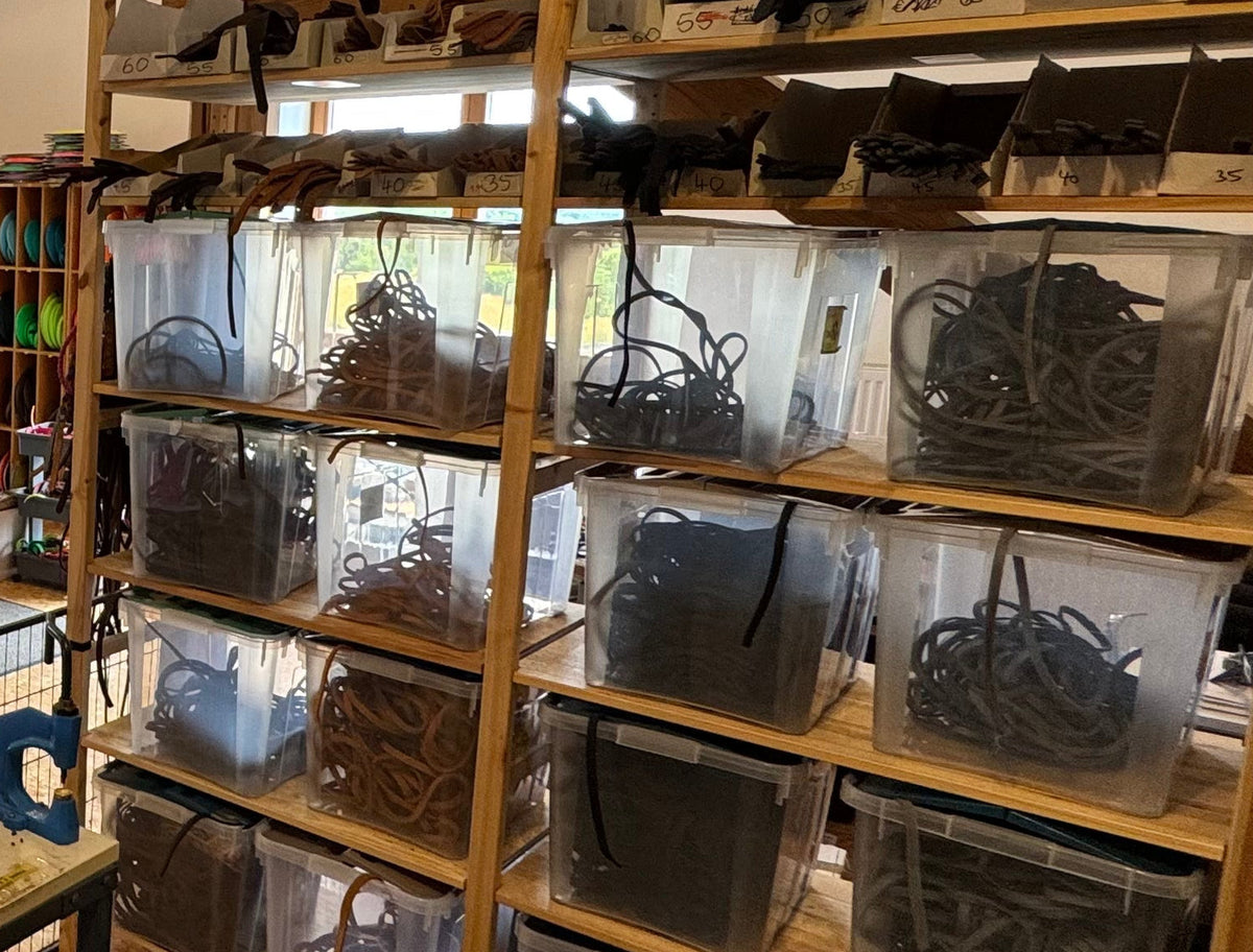 Materiallager mit aufgerollten Leder- und Biothane-Riemen in beschrifteten Boxen auf der Regalwand der Werkstatt